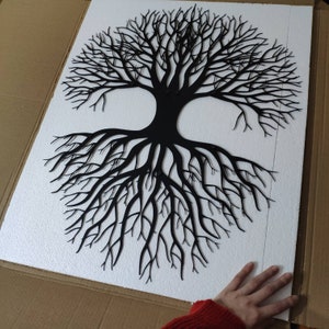 May include: Black metal tree of life wall art. The tree design features a detailed depiction of branches and roots. The artwork is mounted on a white surface, creating a striking contrast. The piece is likely for home decor.
