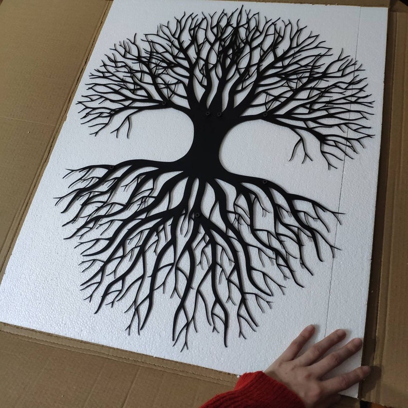 May include: Black metal tree of life wall art. The tree design features a detailed depiction of branches and roots. The artwork is mounted on a white surface, creating a striking contrast. The piece is likely for home decor.