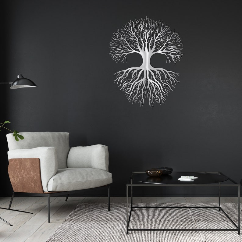 May include: A silver metal tree of life wall art decor. The tree design features a symmetrical arrangement of branches and roots. The artwork is mounted on a dark gray wall, creating a striking contrast. The room also includes a modern armchair, a coffee table, and a rug.