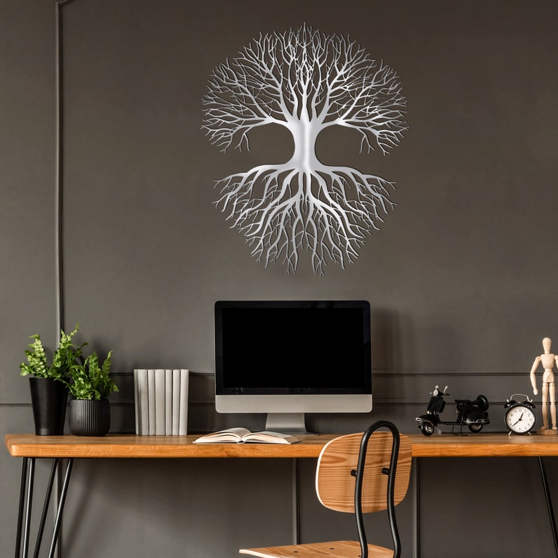 May include: A silver metal tree of life wall art decor. The tree design features a symmetrical arrangement of branches and roots. The artwork is mounted on a dark gray wall above a wooden desk with a computer and other decorative items.