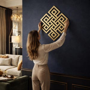 Op de afbeelding: Een goudkleurig, geometrisch wandkunstwerk wordt op een donkerblauwe muur gemonteerd. Het kunstwerk heeft een ingewikkeld, symmetrisch ontwerp. De persoon op de foto draagt een witte blouse met kleine zwarte stippen en een beige broek.