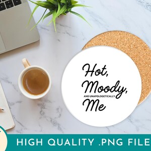 May include: A white ceramic coaster with the text "Hot, Moody, and Unapologetically Me" in black script. The coaster sits on a cork base. A cup of coffee, a laptop, a notepad, and a pencil are also visible.