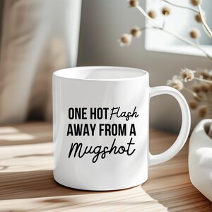 May include: White ceramic mug with the text "ONE HOT FLASH AWAY FROM A Mugshot" in black. The mug has a curved handle and is sitting on a wooden surface.