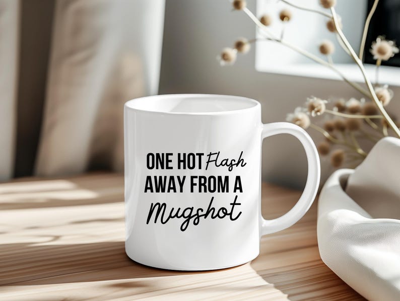 May include: White ceramic mug with the text "ONE HOT FLASH AWAY FROM A Mugshot" in black. The mug has a curved handle and is sitting on a wooden surface.