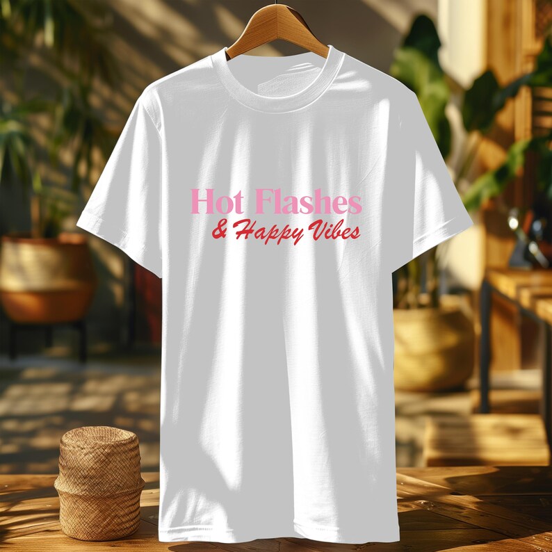 May include: White t-shirt with the text "Hot Flashes & Happy Vibes" in pink script. The shirt is displayed on a wooden hanger against a blurred background of plants and furniture. The t-shirt is a crew neck style.