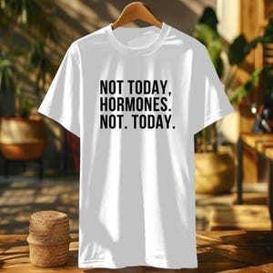 May include: A white t-shirt with the text "NOT TODAY, HORMONES. NOT. TODAY." printed in black. The shirt is hanging on a wooden hanger. The background is blurred, showing a glimpse of a wooden table and plants.