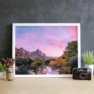 May include: A framed photograph of a mountain range with a river flowing through the valley. The sky is a vibrant pink and purple, with clouds scattered across the horizon. The mountains are a muted brown and green, with trees lining the riverbanks.