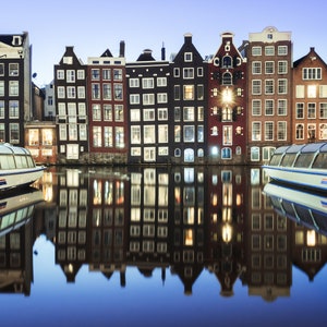 Puede incluir: Una vista de un canal en Ámsterdam, Países Bajos, al atardecer. El canal está bordeado de edificios tradicionales de ladrillo con muchas ventanas. Dos barcos-canal blancos están atracados a ambos lados del canal. Los barcos tienen la palabra "plas" en el lateral.