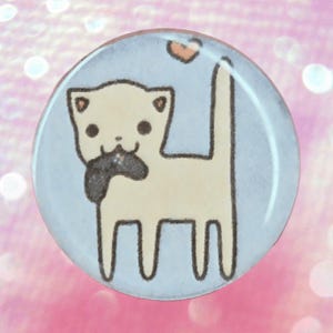 Op de afbeelding: Een ronde knoop met een lichtblauwe achtergrond met een cartoonkat die een videogamecontroller vasthoudt. De kat is crèmekleurig met zwarte contouren en een klein hartje boven zijn staart.