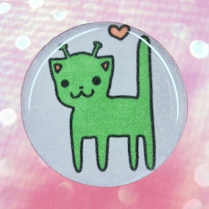 May include: A round button with a cartoon alien cat design. The alien cat is green with black outlines, a small orange heart, and a white background. The button has a glossy finish.
