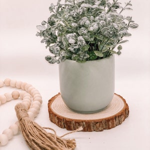 Pode incluir: Um pequeno vaso de cer&acirc;mica verde claro cont&eacute;m uma planta artificial com folhas verdes empoeiradas. O vaso assenta sobre um porta-copos redondo de fatia de madeira natural. Uma fileira de contas de madeira e uma borla de juta est&atilde;o em primeiro plano.