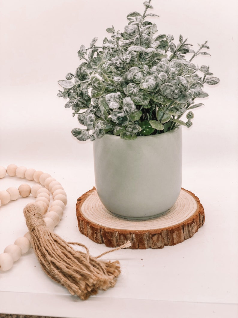 Pode incluir: Um pequeno vaso de cer&acirc;mica verde claro cont&eacute;m uma planta artificial com folhas verdes empoeiradas. O vaso assenta sobre um porta-copos redondo de fatia de madeira natural. Uma fileira de contas de madeira e uma borla de juta est&atilde;o em primeiro plano.