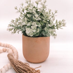 Pode incluir: Uma planta de eucalipto artificial com folhas verdes e brancas num vaso de cer&acirc;mica cor de p&ecirc;ssego. Uma grinalda de contas de madeira com uma borla de juta est&aacute; em primeiro plano. Decora&ccedil;&atilde;o de casa.