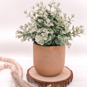 Pode incluir: Um pequeno vaso redondo de terracota cheio de vegeta&ccedil;&atilde;o de eucalipto artificial. O vaso est&aacute; sobre uma fatia de madeira natural, com um fio de contas de madeira e uma borla em primeiro plano. As folhas s&atilde;o verdes e brancas.