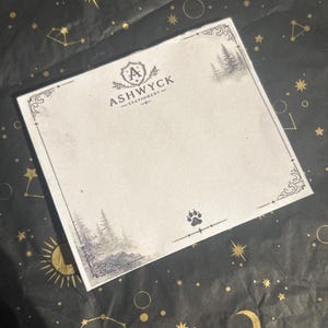 May include: A square sheet of off-white stationery paper featuring a dark gray border with decorative corners. The top center displays a shield logo with the letter A and the text ASHWYCK STATIONERY. A forest illustration and a paw print are visible.