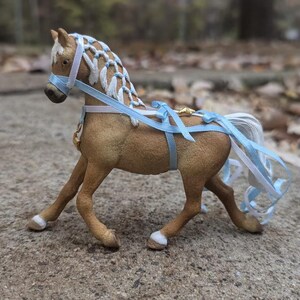 May include: A light brown horse figurine with a white mane and tail, wearing a blue ribbon halter and a blue ribbon tied around its neck and tail.