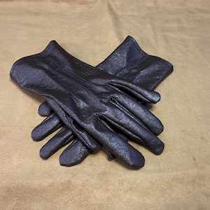 May include: A pair of dark brown leather gloves, with a textured surface, are crossed over each other. The gloves have a classic design with visible stitching and are set against a tan background.