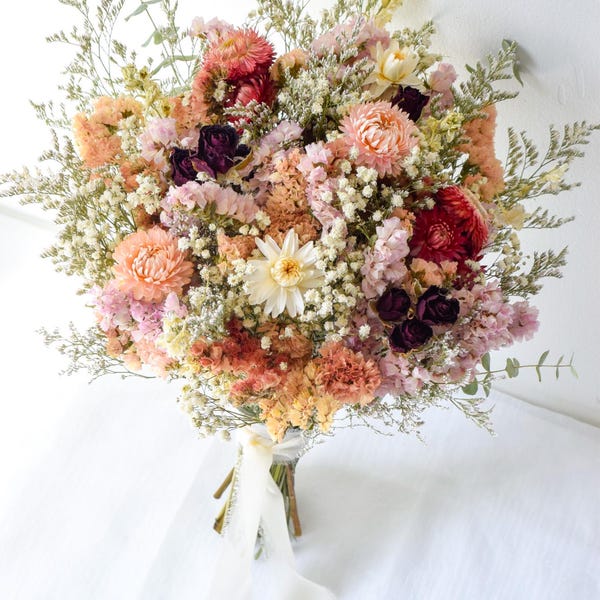 May include: A bouquet of dried flowers featuring shades of peach, pink, cream, and deep burgundy. The arrangement includes strawflowers, baby's breath, and greenery, tied together at the base with a long, flowing white ribbon.