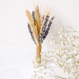 May include: A small bouquet of dried lavender and wheat tied together with twine. The bouquet is on a white background with white baby's breath flowers in the foreground.
