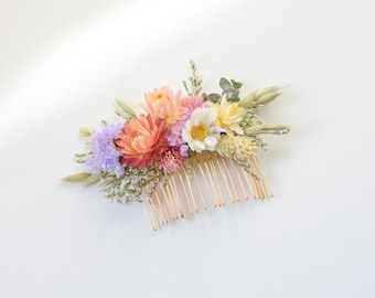May include: A floral hair comb featuring a variety of dried flowers in shades of peach, pink, yellow, and lavender. The flowers are arranged on a gold-colored metal comb, perfect for weddings or special occasions.