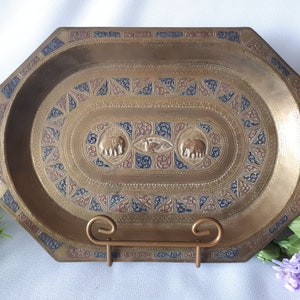 May include: Octagonal brass tray with an oval inset, featuring intricate etched designs in gold, red, and blue. Two elephants are depicted in the centre, with a bird in between them.