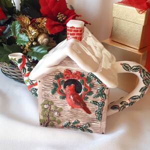 Puede incluir: Una tetera de cerámica con forma de casita de pájaros con un techo nevado, una chimenea de ladrillo rojo y un cardenal posado en una rama. La tetera está decorada con hojas de acebo verdes y bayas rojas.