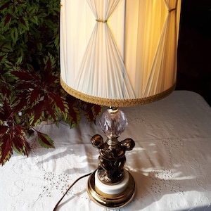 Puede incluir: Una lámpara de mesa vintage con una pantalla de lámpara blanca plisada y una base dorada con dos querubines. La pantalla tiene un ribete dorado y la base tiene un remate de cristal transparente.