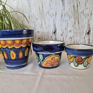 May include: Three ceramic plant pots with blue rims and vibrant patterns. The largest pot features yellow and brown teardrop designs. The other two pots display floral and swirl patterns in orange, green, and blue.