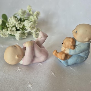 Op de afbeelding: Twee keramische babybeeldjes. Eén ligt in roze, de andere zit en houdt een kleine bruine teddybeer vast. De zittende baby draagt een blauw pak. Witte bloemen staan op de achtergrond.