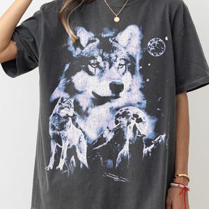 May include: Dark gray t-shirt featuring a detailed graphic of a wolf with a moon and stars. The design includes multiple wolves and a night sky scene. The shirt has short sleeves and a relaxed fit.