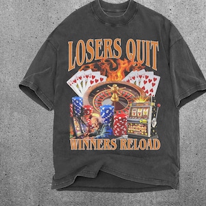 Könnte beinhalten: Schwarzes T-Shirt mit einem grafischen Druck, der eine Casino-Szene mit einem Roulette-Rad, einem Spielautomaten und Spielkarten zeigt. Der Text "Losers Quit Winners Reload" ist auf dem T-Shirt aufgedruckt.