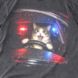 May include: Dark grey t-shirt with a graphic of a cat driving a car, with flashing police lights. The cat has a surprised expression and is holding the steering wheel. The shirt is made of a soft material.