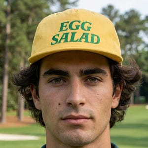 Könnte beinhalten: Gelbe Baseballkappe mit den Worten "EGG SALAD" in grünen Blockbuchstaben. Die Kappe wird von einer Person mit einem grünen Kragenhemd mit grünem und weißem Streifenmuster getragen. Der Hintergrund ist ein Golfplatz.
