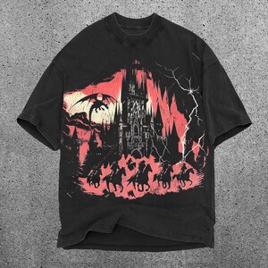May include: Black t-shirt with a graphic print of a castle, a dragon, and people riding horses. The graphic is in shades of red and black.