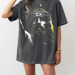 May include: Dark gray t-shirt featuring a graphic of a person with a crown of thorns. The image is in black, white, and yellow. The shirt has short sleeves and a crew neck. The shirt is worn by a person.