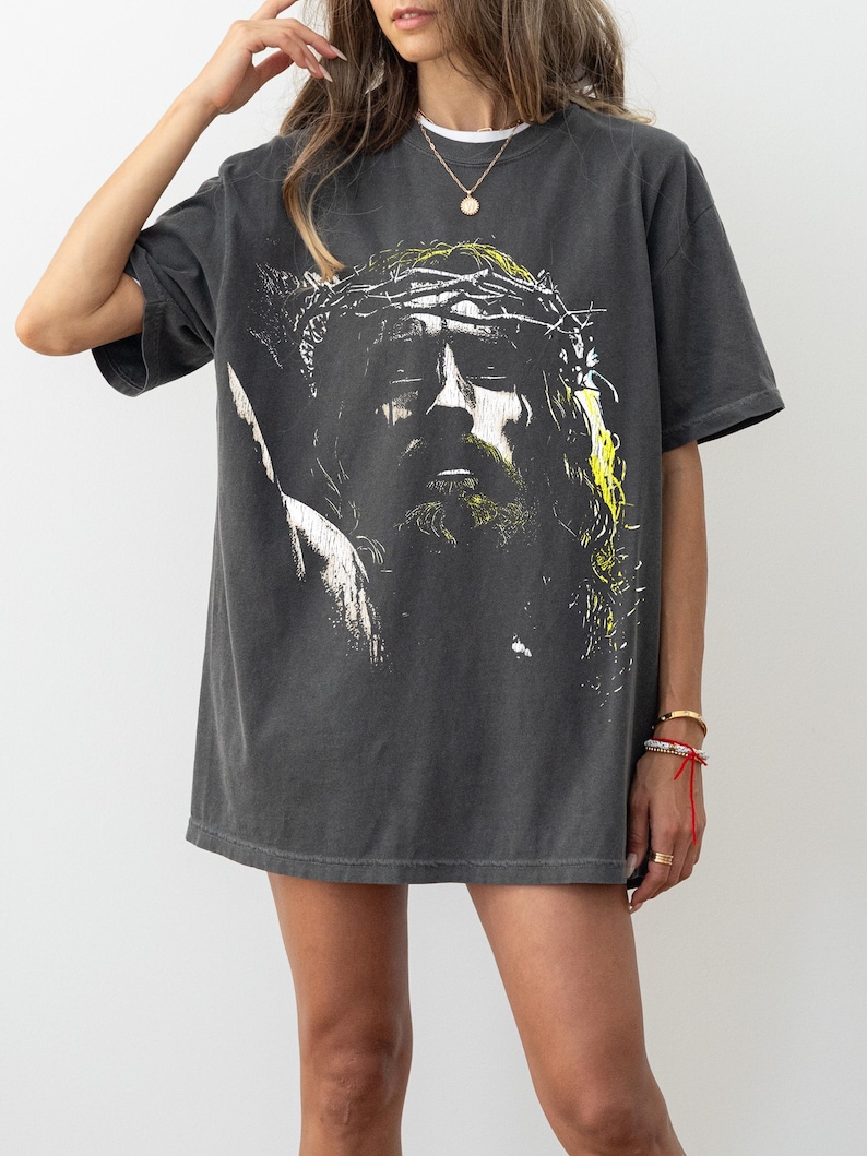 May include: Dark gray t-shirt featuring a graphic of a person with a crown of thorns. The image is in black, white, and yellow. The shirt has short sleeves and a crew neck. The shirt is worn by a person.