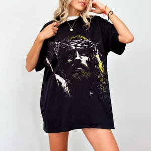 May include: Black graphic t-shirt featuring a detailed illustration of a figure with a crown of thorns. The artwork is predominantly black and white with yellow accents. The shirt has a classic crew neck and short sleeves.