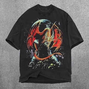 May include: Black t-shirt with a striking graphic of a red and orange dragon with large wings, set against a dark background with a celestial body. The dragon is perched on a rocky surface. The shirt has a classic crew neck.