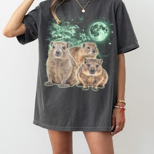 May include: Dark gray t-shirt featuring a graphic of three groundhogs under a glowing green moon and stars. The design has a vintage, distressed look. The shirt is a relaxed fit.