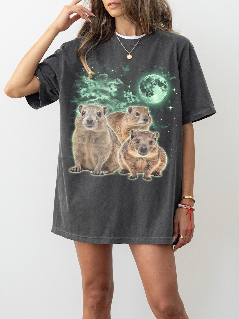 May include: Dark gray t-shirt featuring a graphic of three groundhogs under a glowing green moon and stars. The design has a vintage, distressed look. The shirt is a relaxed fit.