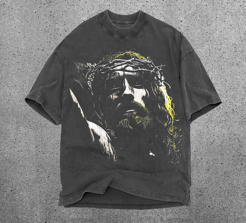 May include: Dark gray t-shirt featuring a distressed graphic of a person wearing a crown of thorns. The graphic is in shades of yellow and white, with a vintage, worn look. The shirt has a crew neck and short sleeves.