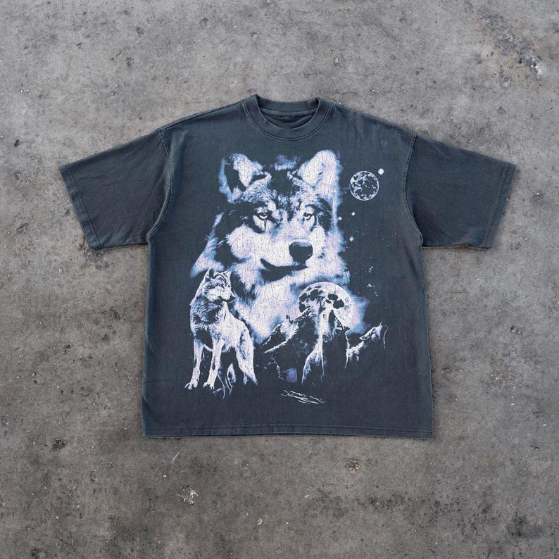 May include: Dark gray t-shirt featuring a detailed white and gray wolf graphic. The design includes a large wolf head, a smaller wolf, a moon, and stars. The shirt has short sleeves and a crew neck.