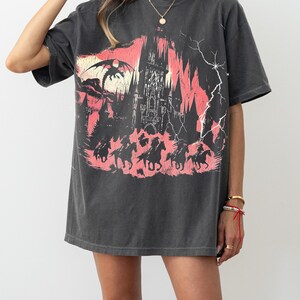 May include: A black oversized t-shirt with a graphic print of a castle, a dragon, and horses. The graphic is in shades of pink and red.