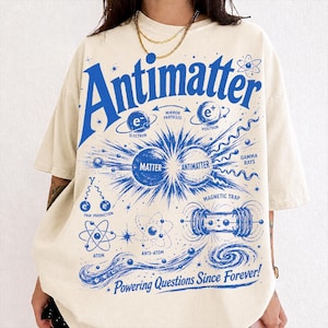 Op de afbeelding: Een beige t-shirt met korte mouwen en een grote blauwe grafische print van wetenschappelijke diagrammen, met de tekst "Antimatter" bovenaan en "Powering Questions Since Forever!" onderaan, met materie, antimaterie en atomen.