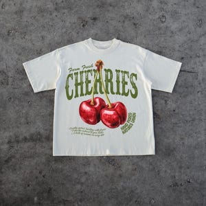 Puede incluir: Una camiseta blanca con un estampado gr&aacute;fico de dos cerezas rojas y el texto "Farm Fresh Cherries Hand Picked Nature's Candy".