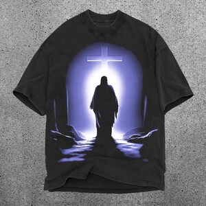 May include: Black t-shirt with a graphic print of a person walking towards a cross in a purple and blue light.