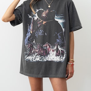 Puede incluir: Camiseta gris oscuro de gran tama&ntilde;o con un gr&aacute;fico de un vaquero con sombrero, con caballos corriendo por el agua. El dise&ntilde;o est&aacute; desgastado, dando a la camiseta un aspecto vintage. La camiseta tiene mangas cortas y cuello redondo.
