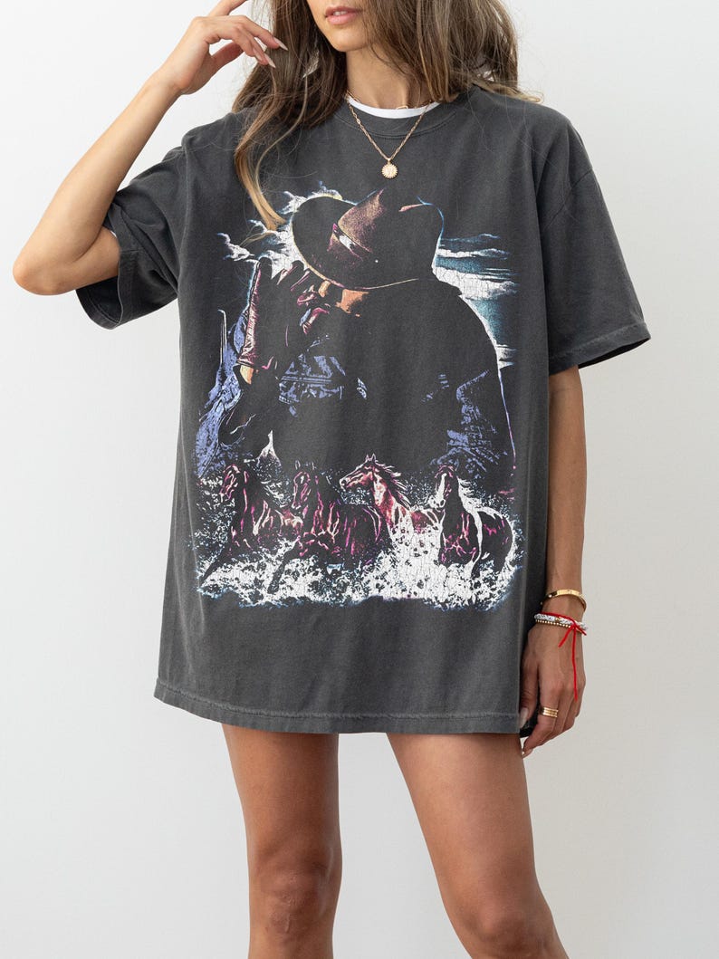 Puede incluir: Camiseta gris oscuro de gran tama&ntilde;o con un gr&aacute;fico de un vaquero con sombrero, con caballos corriendo por el agua. El dise&ntilde;o est&aacute; desgastado, dando a la camiseta un aspecto vintage. La camiseta tiene mangas cortas y cuello redondo.