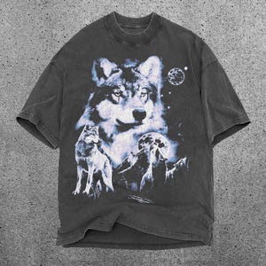 May include: Dark gray t-shirt featuring a detailed wolf graphic. The design includes a large wolf head, a smaller wolf, a full moon, and a starry night sky. The shirt has a crew neck and short sleeves.