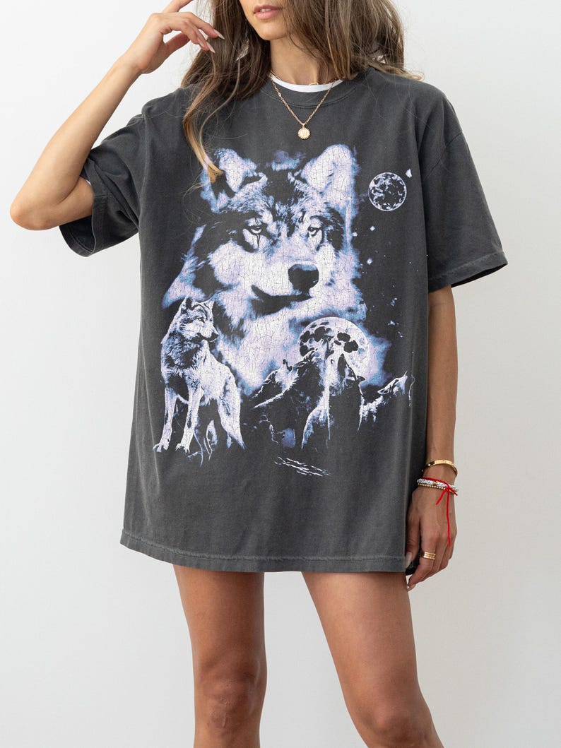 May include: Dark gray t-shirt featuring a detailed graphic of a wolf with a moon and stars. The design includes multiple wolves and a night sky scene. The shirt has short sleeves and a relaxed fit.